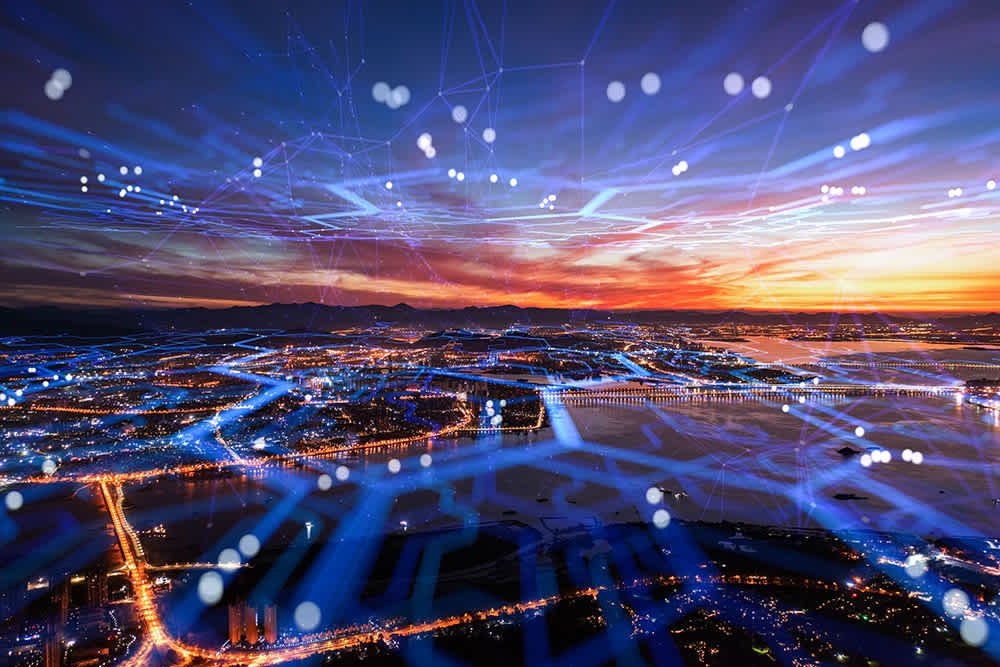A cityscape at sunset with illuminated roads and buildings, overlaid with blue digital network lines and glowing nodes, symbolizing technology and connectivity.