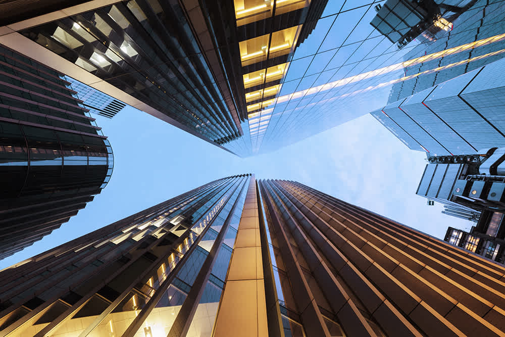 Tall modern skyscrapers viewed from below, converging towards a blue sky. The buildings have reflective glass and illuminated windows, creating dramatic lines and symmetry.