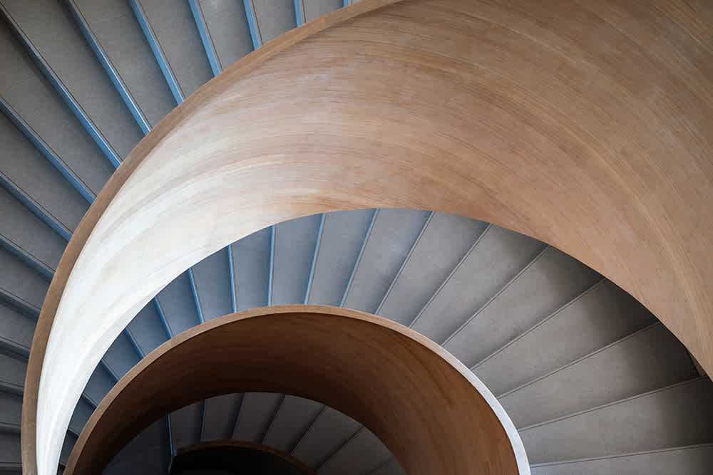 A top-down view of a modern spiral staircase with smooth wooden railings and gray steps, creating an abstract geometric pattern with curves and lines.