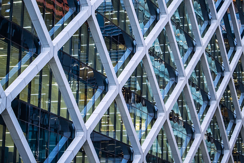 Close-up of a modern building facade with a geometric, crisscrossing steel framework and reflective glass windows, showing the interior lights and reflections of surrounding structures.