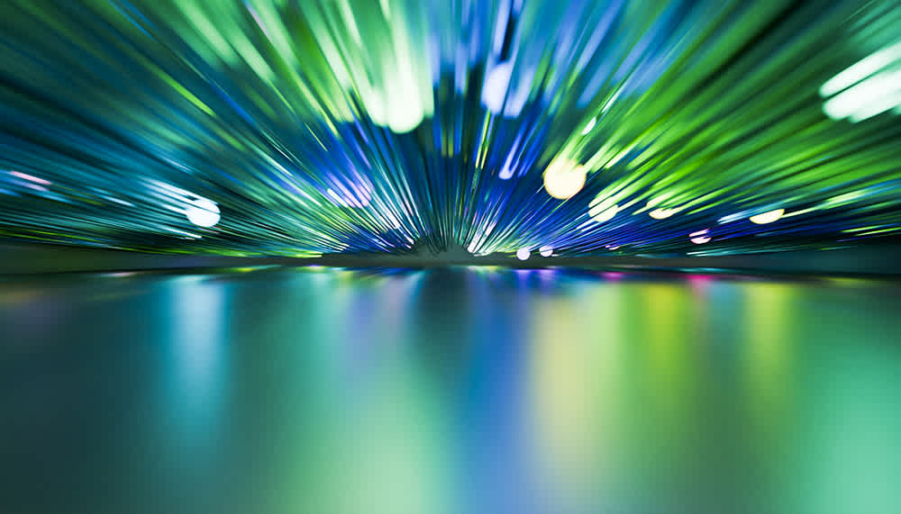 Abstract image of green and blue light streaks radiating outward toward the viewer, resembling a burst of energy or fiber optic cables, with reflections on a smooth, glossy surface below.