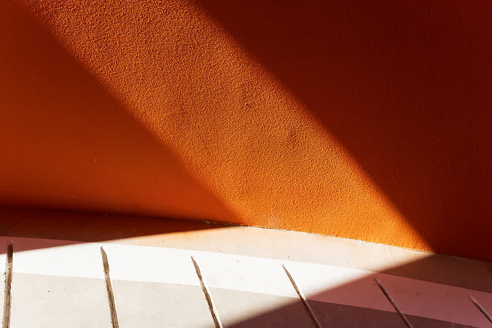 Bright sunlight casts angular shadows on an orange textured wall and a curved, light-colored floor with parallel lines, creating a bold geometric pattern.