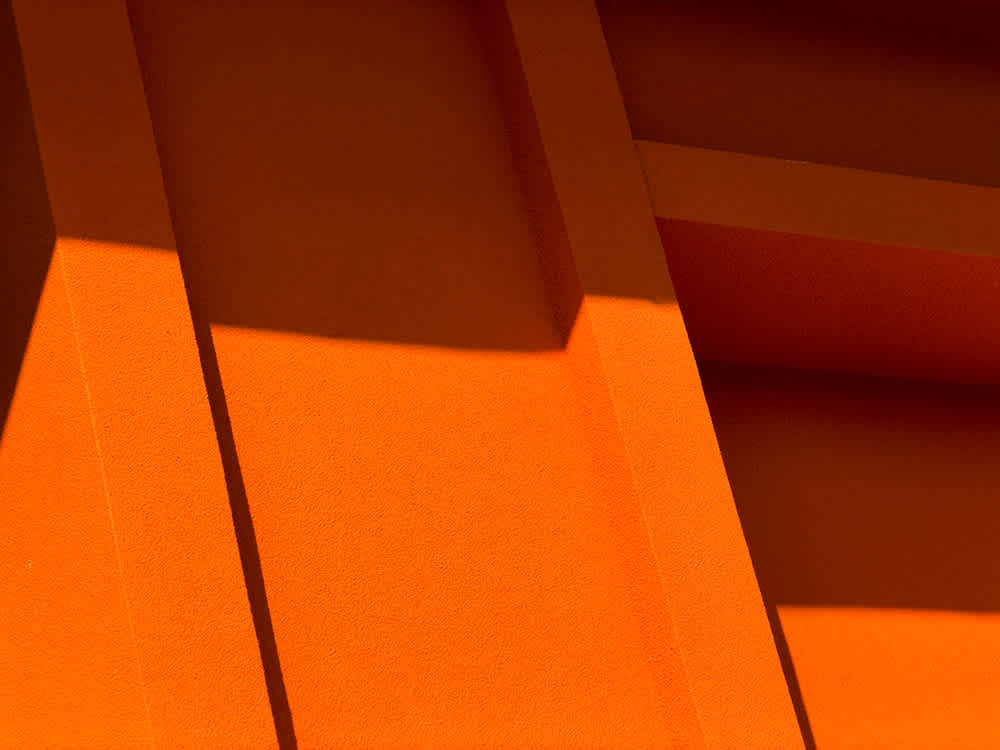 Abstract image showing geometric shapes and shadows on an orange textured surface, with diagonal lines and rectangular panels creating a bold visual pattern.