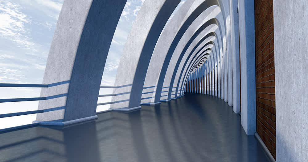 A modern, curved walkway with tall, arched concrete supports and metal railings, casting shadows on the shiny floor. The corridor is open on one side, with a brick wall on the right, and a bright sky visible outside.