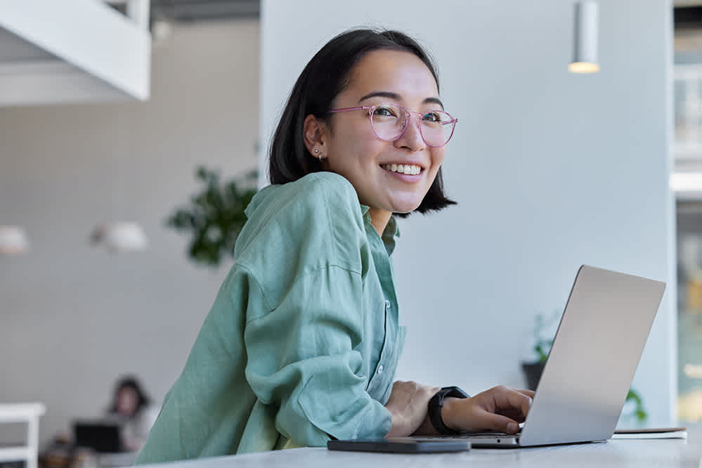 A young woman with glasses and a green shirt smiles while working on a laptop at a bright, modern workspace.