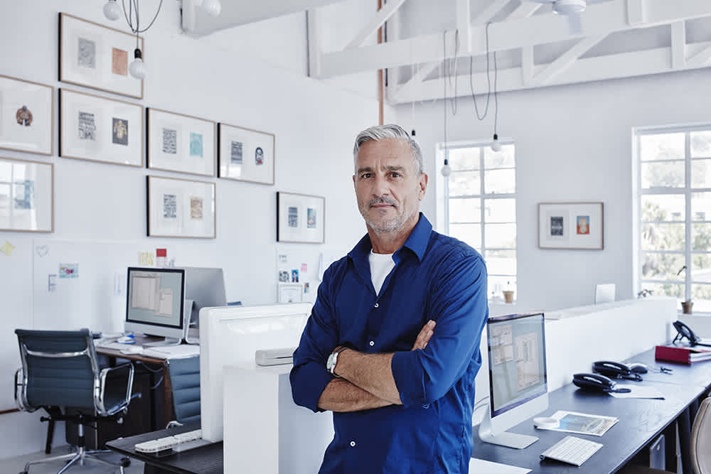 A man with gray hair and a beard, wearing a blue shirt, stands with folded arms in a modern, bright office with desks, computers, and framed art on the white walls.