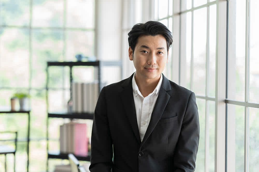 A man in a black suit and white shirt stands by large windows in a bright, modern office. Shelves with books and plants are visible in the background. He is smiling slightly at the camera.