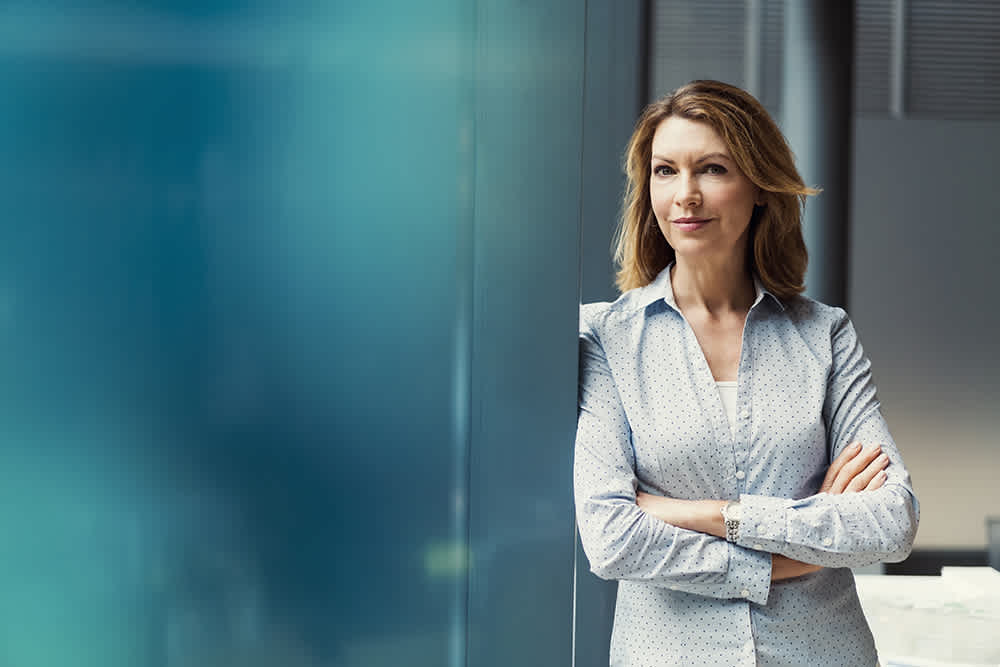 A confident woman with shoulder-length hair stands indoors, leaning against a blue wall with arms crossed, wearing a light blue button-up shirt and smiling slightly. The background is softly blurred.