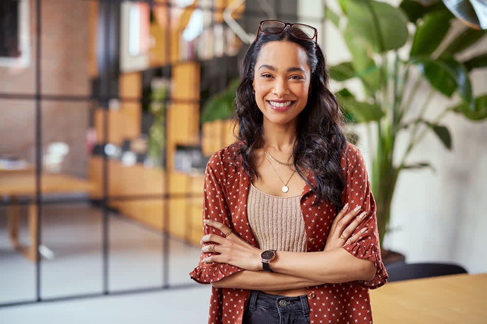A woman with long, dark hair and glasses on her head stands indoors with arms crossed, smiling. She wears a red patterned blouse, a beige top, layered necklaces, and a watch. Modern office setting with plants in the background.