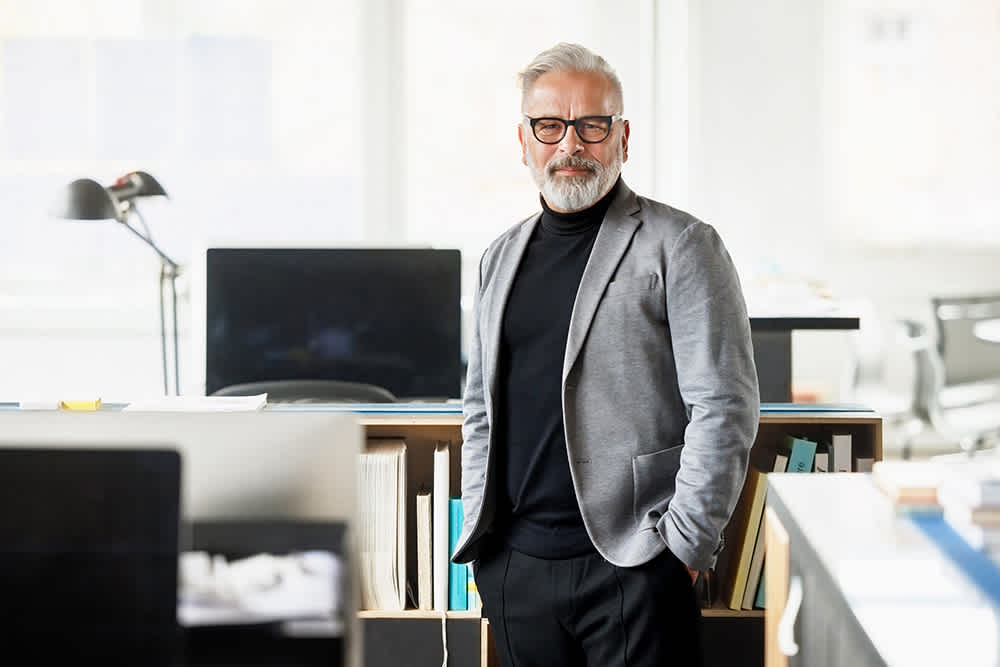 An older man with gray hair, a beard, and glasses stands confidently in a modern office, wearing a gray blazer and black turtleneck, with shelves, desks, and computers in the background.