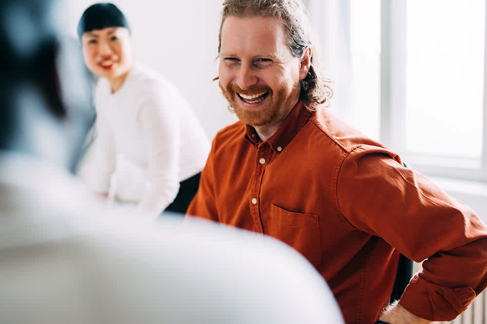 A man with long hair and a beard, wearing an orange shirt, smiles and laughs while sitting in a bright room. A woman in a white top is in the background, also smiling.