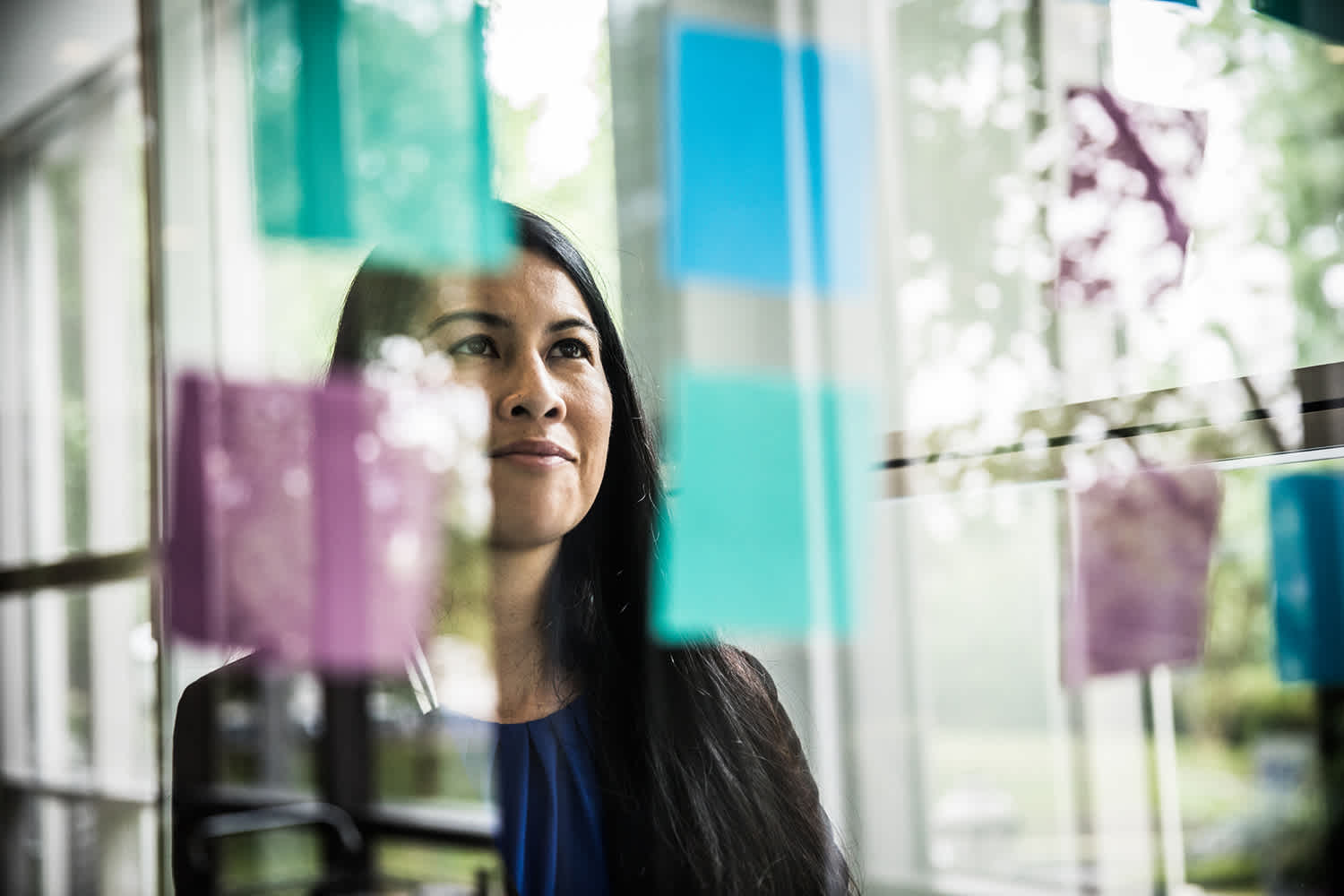 A woman stands indoors, looking thoughtfully at colorful sticky notes on a glass wall with trees visible outside through the windows.