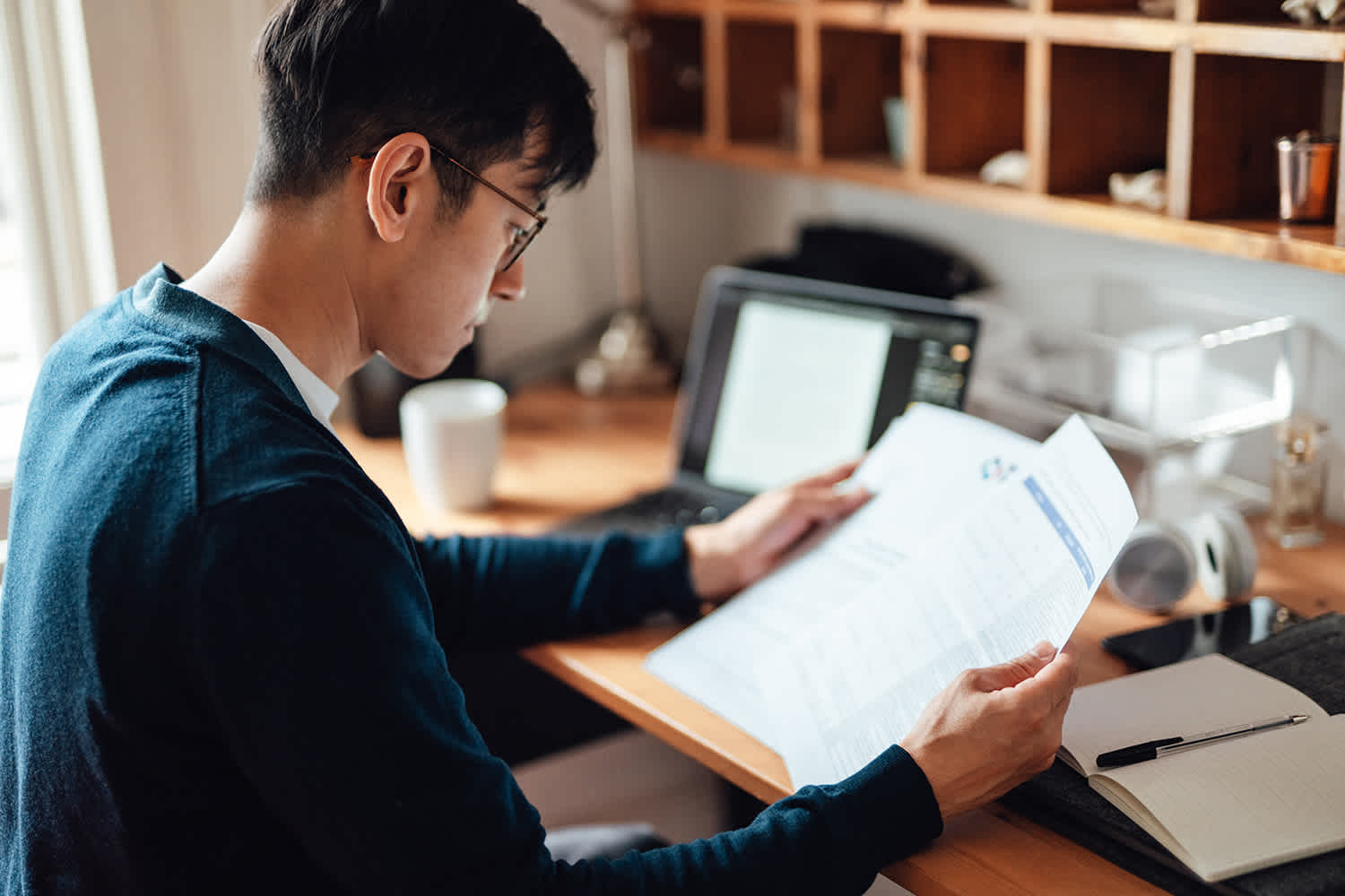 A person wearing glasses and a dark sweater sits at a desk, reading a document. A laptop, notebook, mug, and shelves with small items are visible in the background. The setting appears to be a home office.