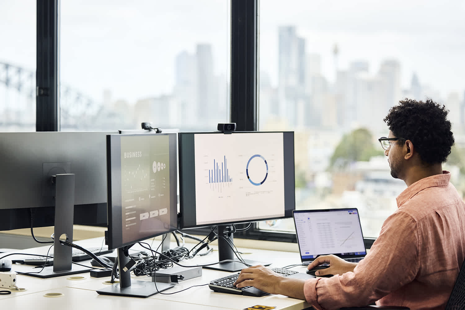 A man sits at a desk in an office with large windows, working on a laptop and two monitors displaying business charts and graphs. A city skyline is visible in the background.