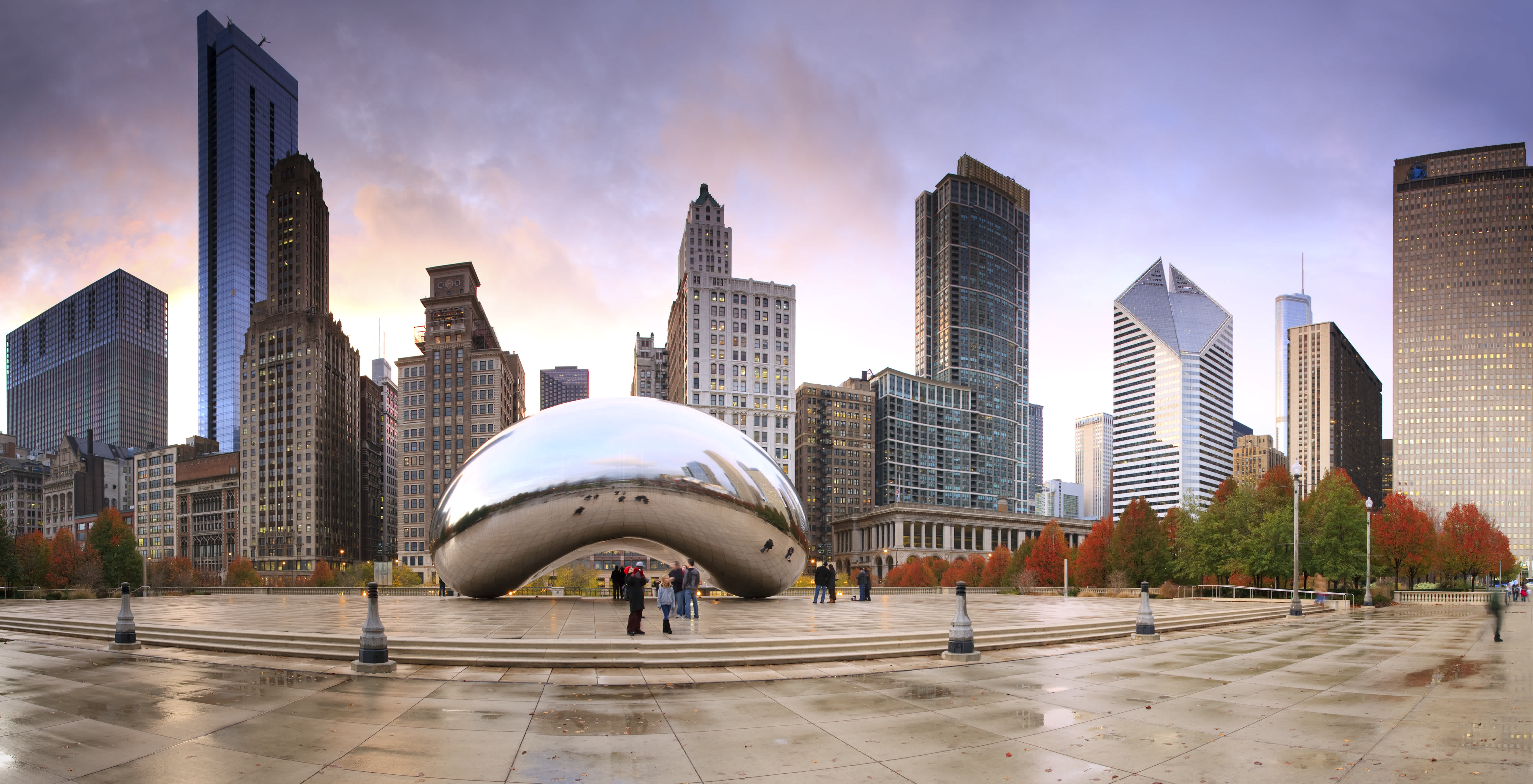 Cloud Gate (The Bean) sculpture in Chicago’s Millennium Park reflects surrounding skyscrapers and colorful autumn trees under a cloudy sky, with a few people walking nearby on wet pavement.