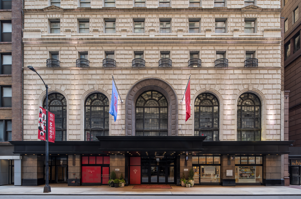 Historic stone building with tall arched windows, three flagpoles with flags above a black awning, and a clean sidewalk in front. The building has detailed stonework and multiple upper-floor windows.