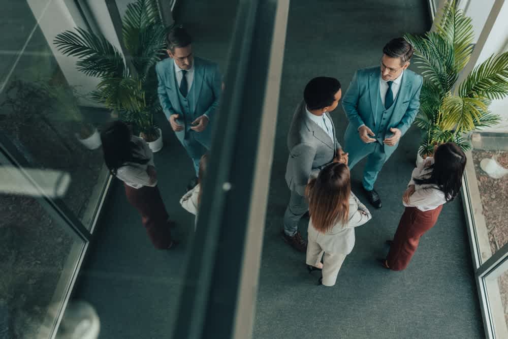 Four professionals stand in a small group, engaged in conversation in an office hallway with large windows and potted plants. The image is taken from above, showing their reflection on the glass wall.