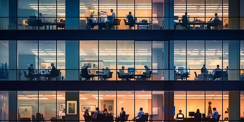 A view of a modern office building at night, showing several floors through large glass windows. People are working at desks, having meetings, and using computers in brightly lit rooms.