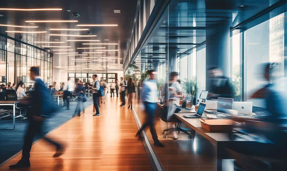 A busy modern office with people walking and working at desks. Large windows line the walls, flooding the space with natural light. The interior features glass partitions and contemporary furniture.