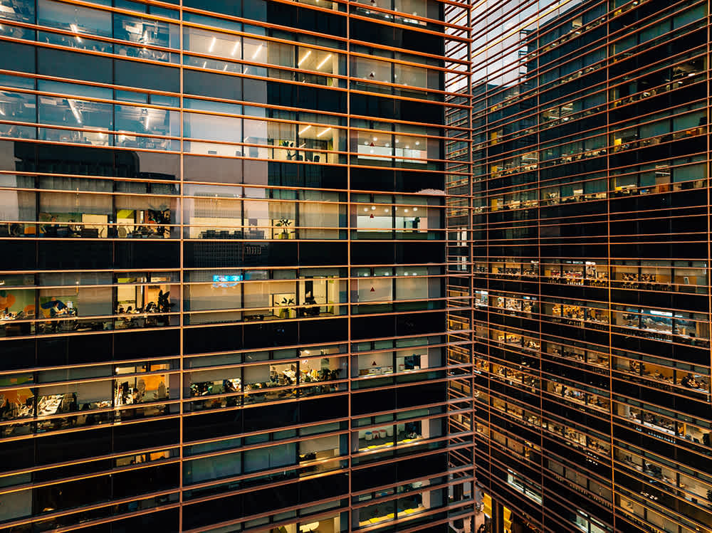 View of two modern office buildings at night, with many illuminated windows revealing people working inside, creating a grid-like pattern of lights and reflections.
