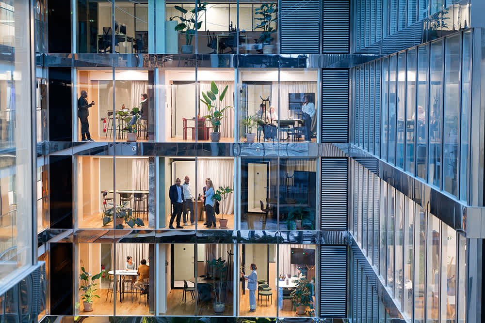 View through the glass facade of a modern office building, showing multiple floors with people working, meeting, and talking in brightly lit rooms filled with plants and contemporary furniture.