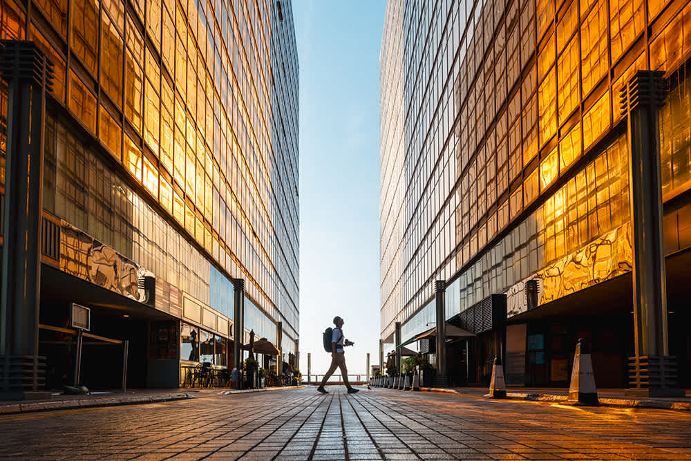 A person with a backpack walks between two tall, glass buildings reflecting golden sunlight, with a blue sky above and brick pavement below. The scene is symmetrical and urban.