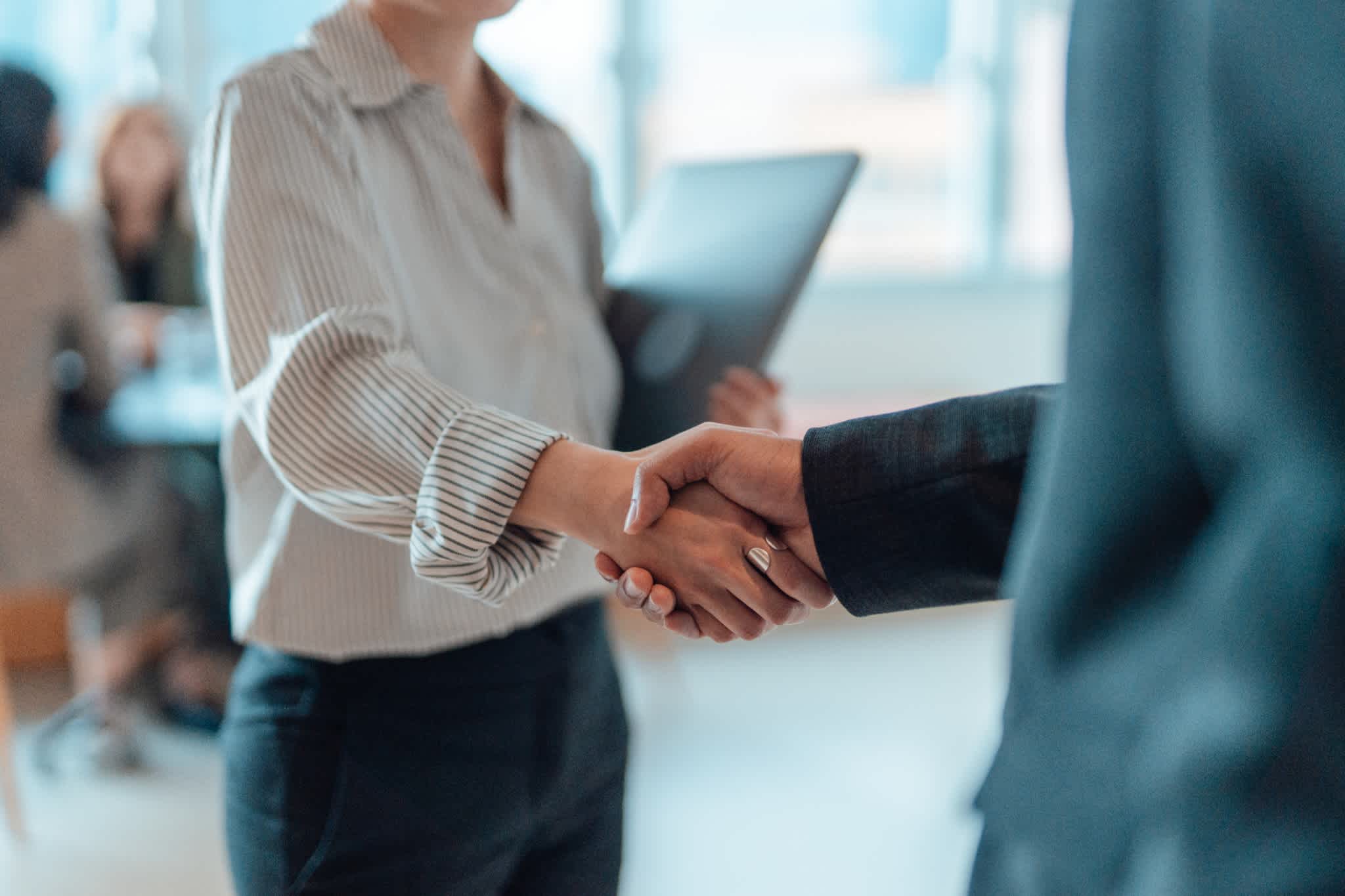 Two people in business attire shake hands indoors, suggesting a professional agreement or introduction, with one person holding a folder and blurred colleagues in the background.
