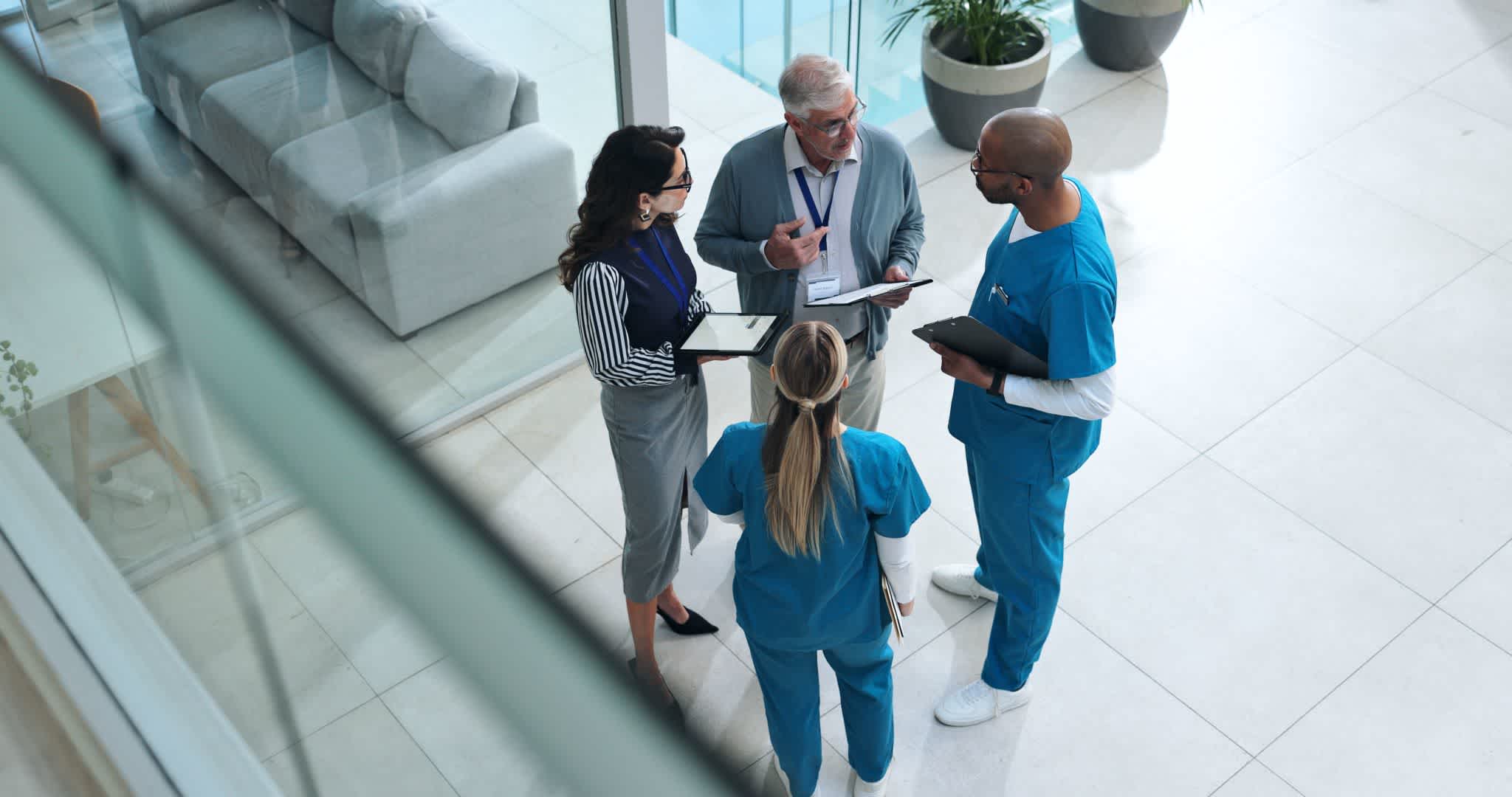 Four professionals, including two people in blue medical scrubs, one in business attire, and an older man with a lanyard, stand in a bright lobby having a discussion and holding tablets or clipboards.