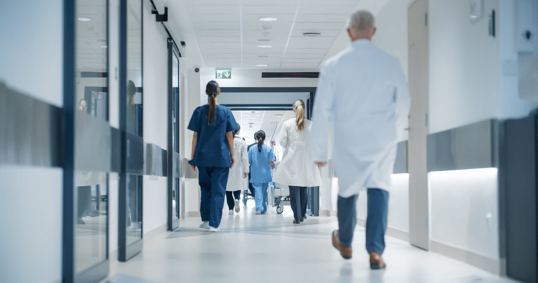 Doctors and nurses in scrubs and lab coats walk down a bright hospital corridor, heading towards patient rooms and work areas. The hallway is clean and modern, with medical equipment visible in the distance.