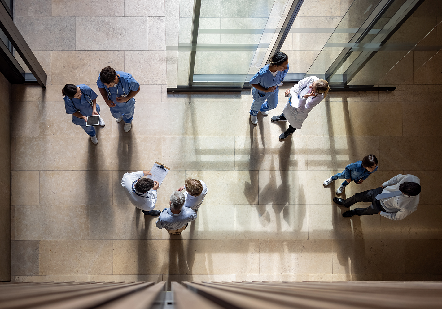 Overhead view of people in medical scrubs and business attire walking and talking in a spacious, sunlit hallway with large glass doors and tiled flooring.