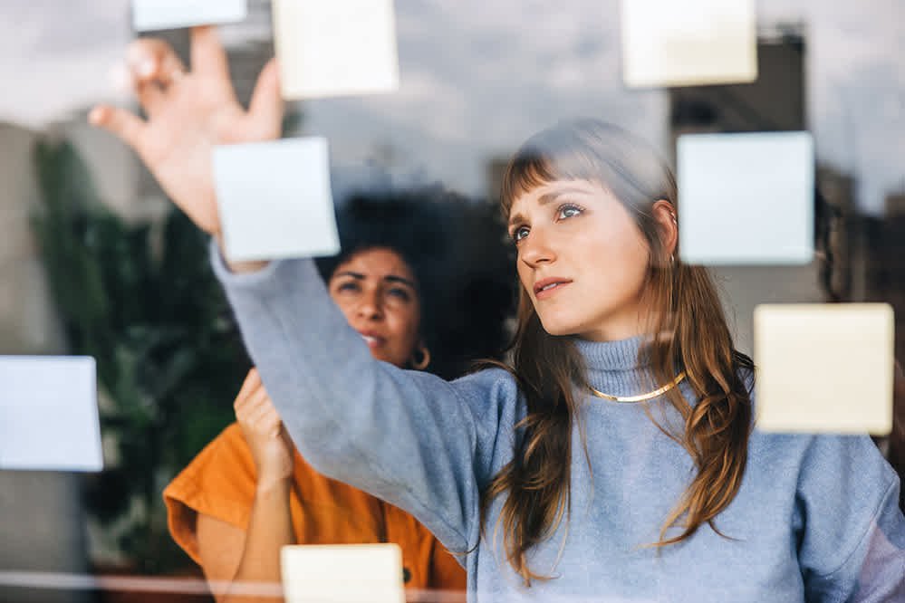 Two women are seen through a window, placing or organizing sticky notes on the glass. One woman in a blue sweater is reaching up, while the other in an orange top looks on, both focused on the notes.