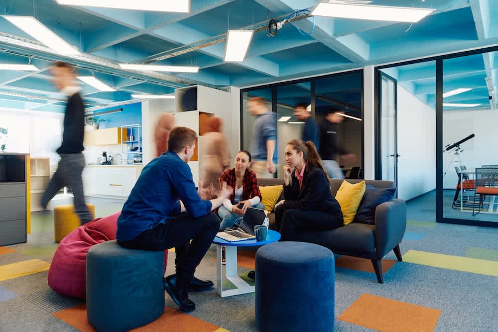 Three people sit talking around a coffee table in a modern office lounge, while several others walk by in the background. The space has colorful decor and glass-walled meeting rooms.
