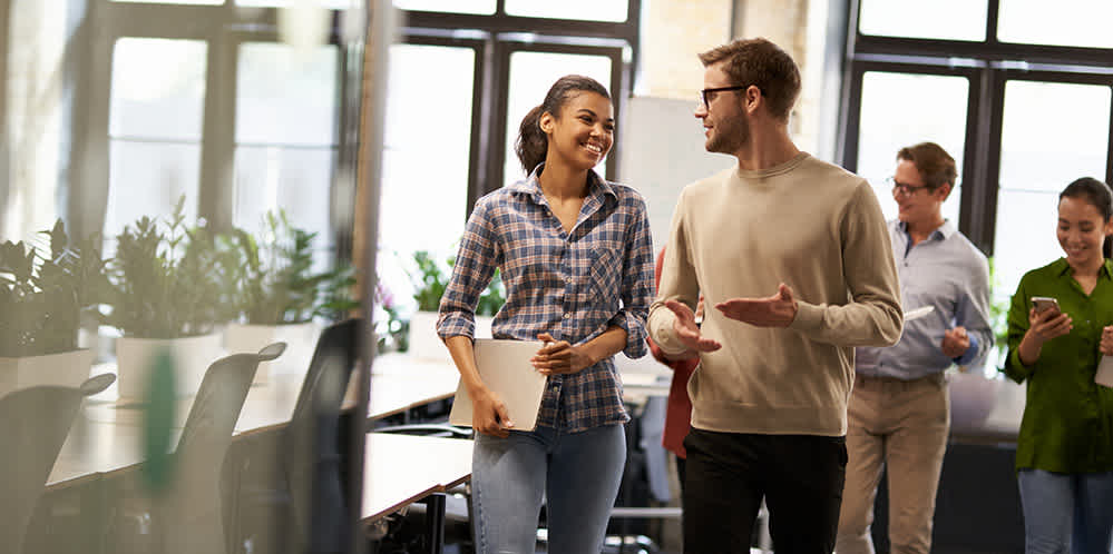 A group of four people, two men and two women, walk together in a bright office space. Two are conversing and smiling, while the others follow behind; one uses a phone. Desks, chairs, and plants are visible in the background.