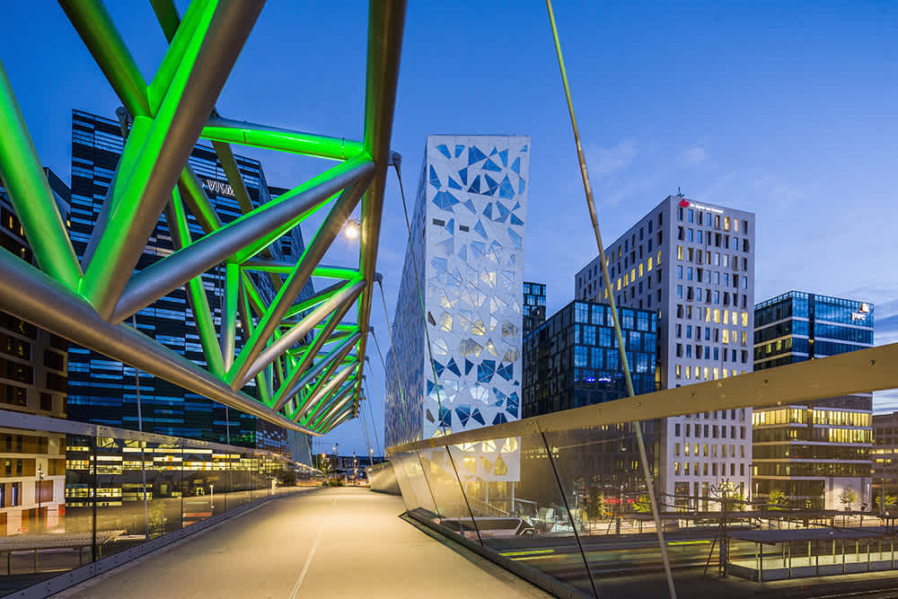 Modern pedestrian bridge with green-lit metal beams spans over a walkway, surrounded by contemporary glass and geometric-patterned office buildings at dusk in an urban cityscape.