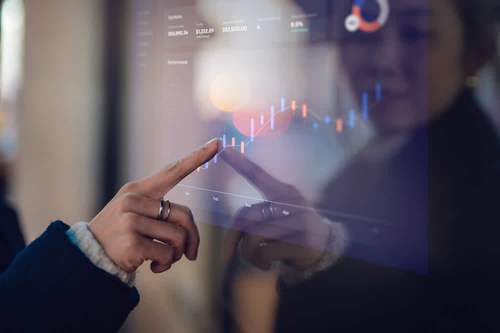 A person touches a digital screen displaying financial graphs and data, including a colorful line chart, portfolio balance, and performance metrics. Their reflection is visible on the screen.