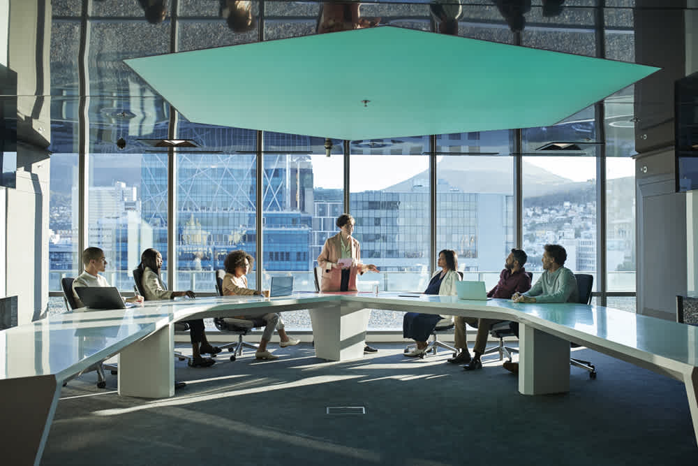 A group of people sit around a modern, U-shaped conference table in a glass-walled office with city buildings visible outside. One person stands and speaks while the others listen attentively.