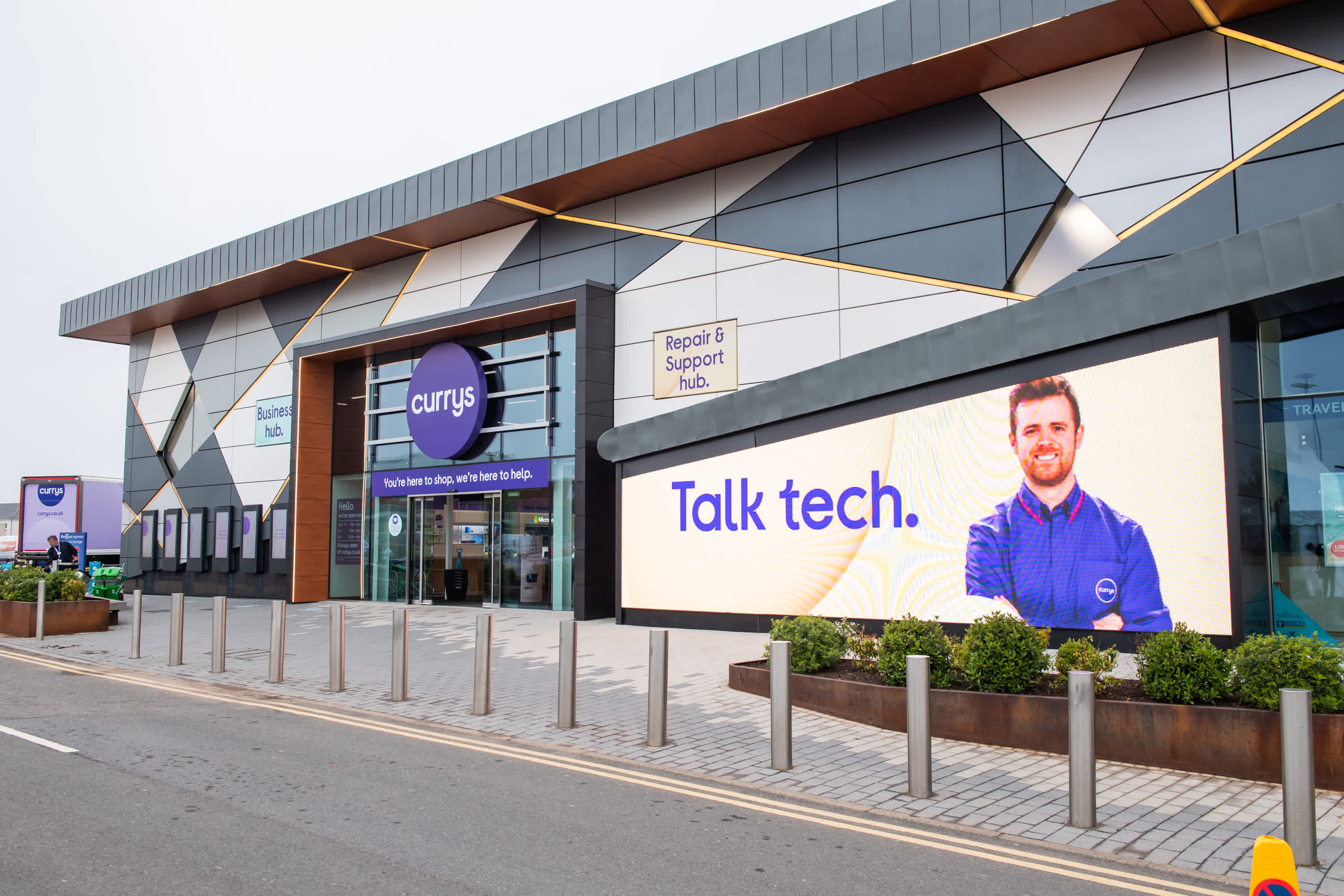 Large Currys electronics store exterior with a modern design. A digital billboard displays a man in a blue shirt and the words Talk tech. The entrance and signs for a repair and support hub are visible.