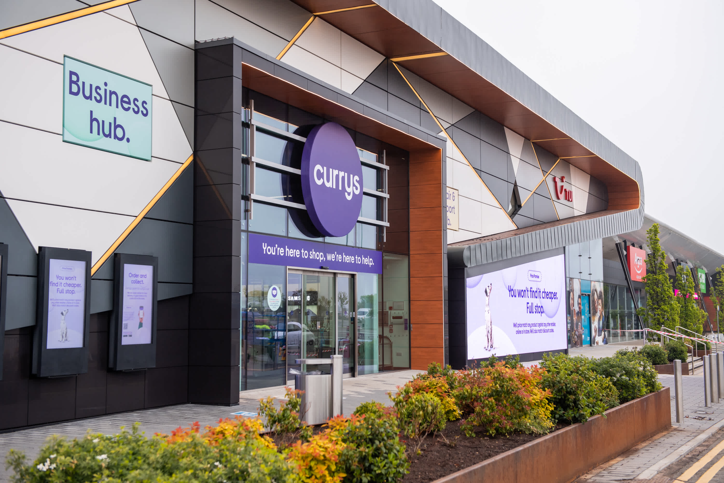 A modern retail store exterior with a large “currys” sign above the entrance, surrounded by digital screens and signs reading “Business hub.” The building is sleek, with plants and a walkway in front.