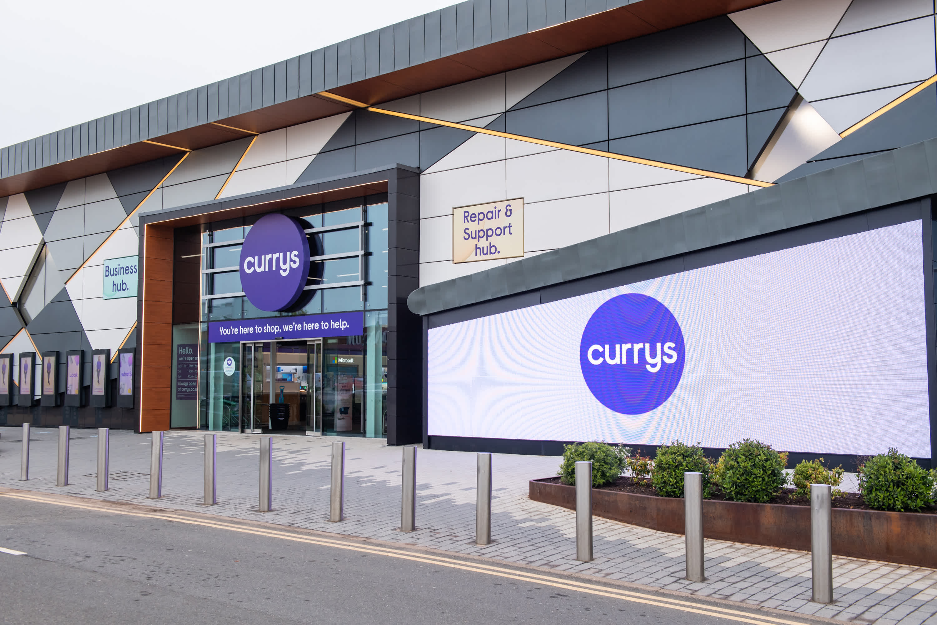 The exterior of a Currys store featuring large glass doors, Currys logos, a digital sign, and signs for the Business hub and Repair & Support hub. Metal bollards line the sidewalk in front.