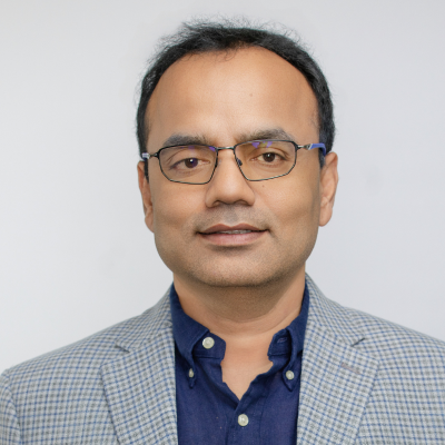 A man with short black hair and glasses, wearing a blue button-up shirt and a gray checkered blazer, poses against a plain light background.