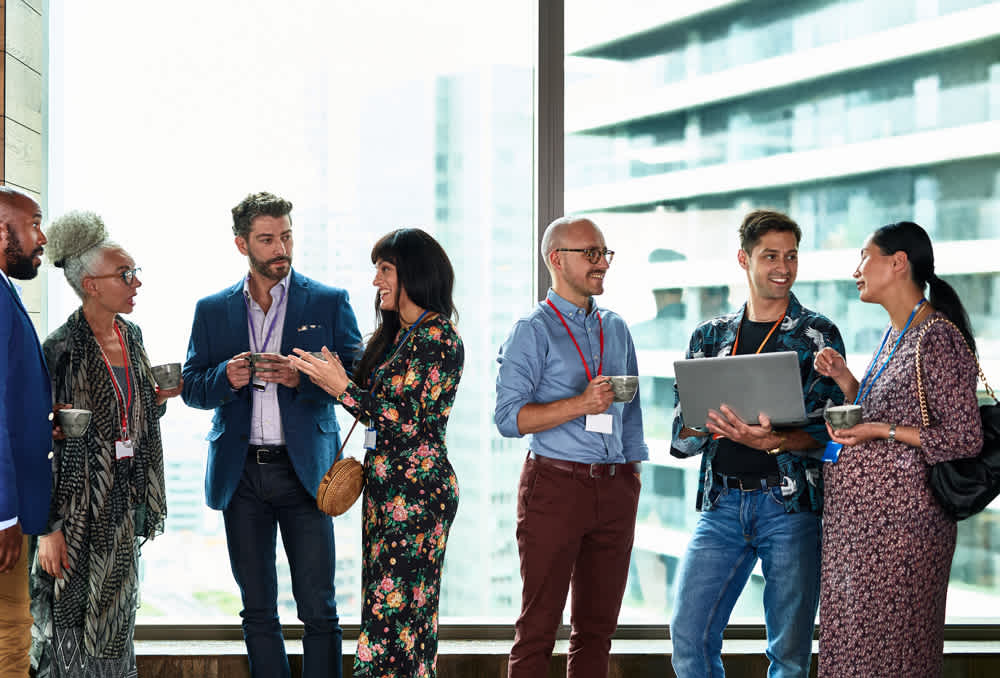 A group of professionally dressed people stand by large windows, talking and holding drinks at a networking event. Two people are looking at a laptop while others chat in small groups.