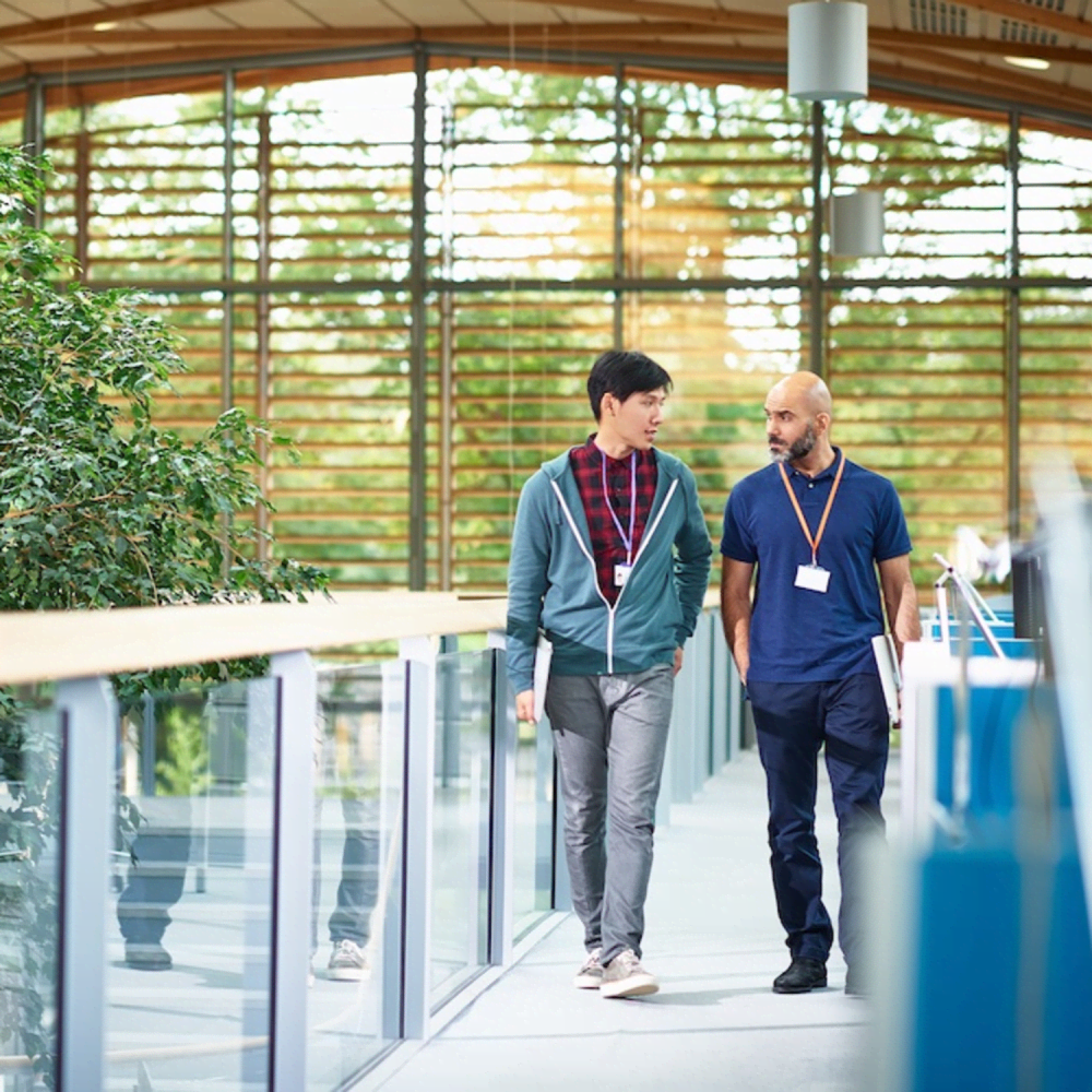 Two people walk side by side through a modern building with glass walls and greenery, engaged in conversation. Both wear ID badges, and natural light streams in from large windows in the background.