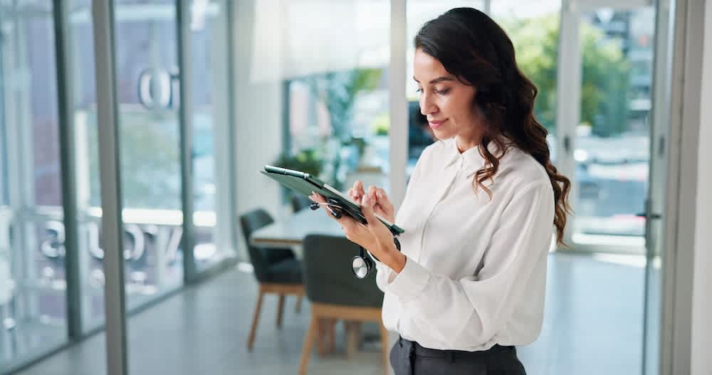 A woman in business attire stands in a modern office, using a tablet. She appears focused, with natural light coming through large windows. Office furniture and greenery are visible in the background.