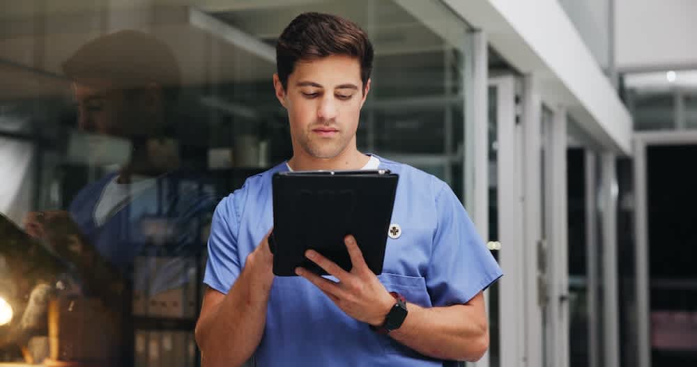 A healthcare professional in blue scrubs stands indoors, looking at and using a tablet device, with glass walls and a reflection visible in the background.