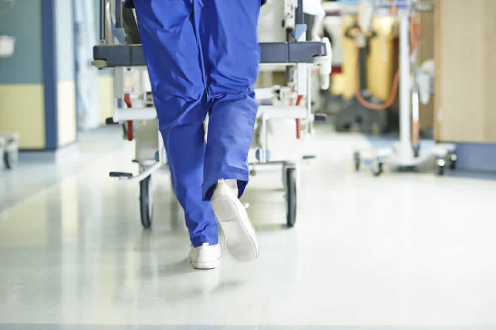 A person in blue medical scrubs and white shoes walks down a hospital hallway, pushing a hospital bed or gurney. The scene appears bright and clinical.