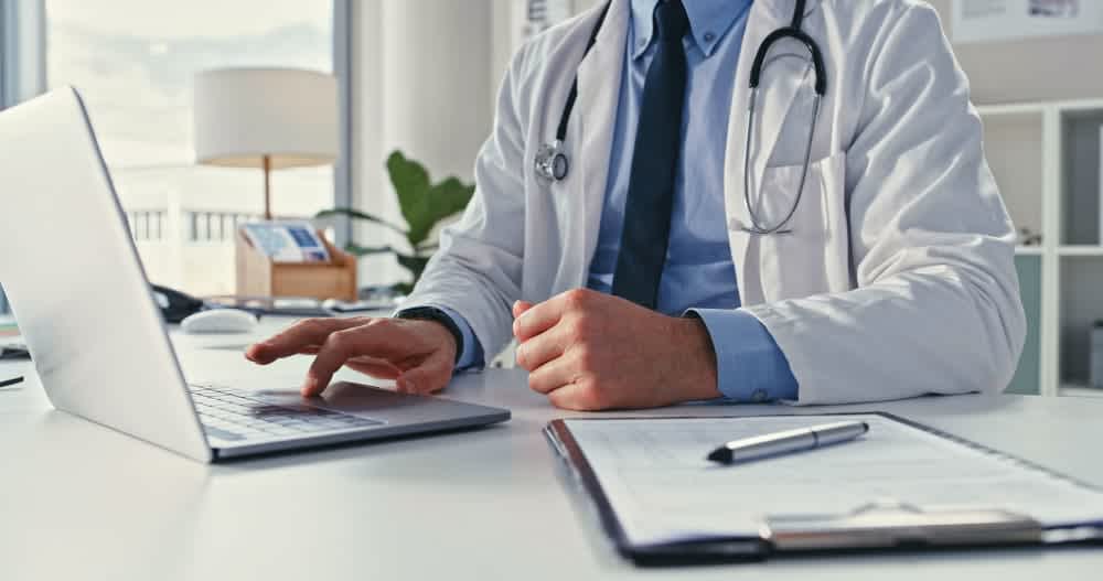 A doctor in a white coat sits at a desk, using a laptop. A stethoscope hangs around their neck, and a clipboard with forms and a pen is on the desk.