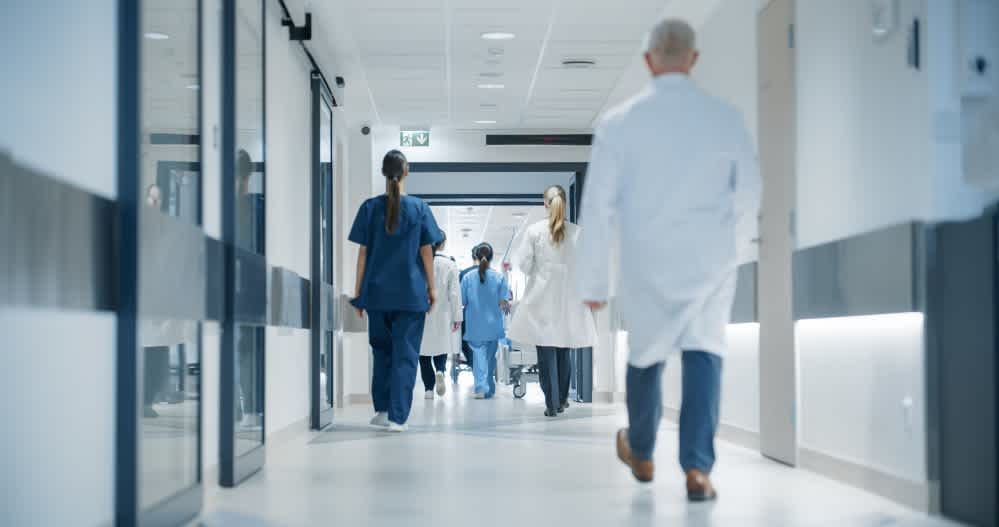 Hospital hallway with medical staff, including doctors and nurses, walking in both directions. The corridor is bright and modern, with glass doors and white walls, creating a clean and professional environment.
