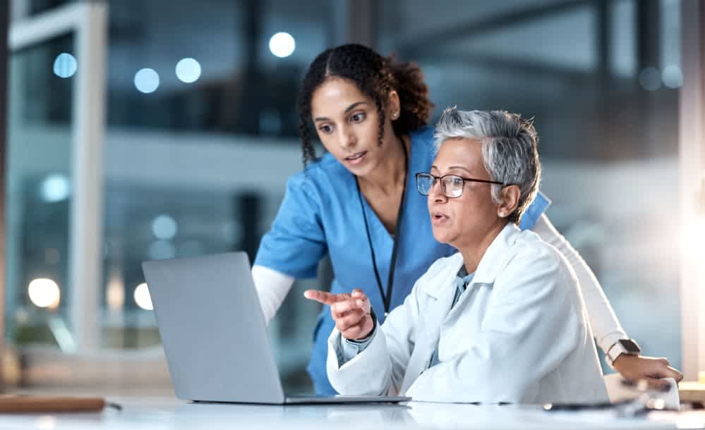 Two healthcare professionals, one in blue scrubs and one in a white coat, discuss information on a laptop in a modern medical office. The person standing looks focused while the seated person gestures at the screen.