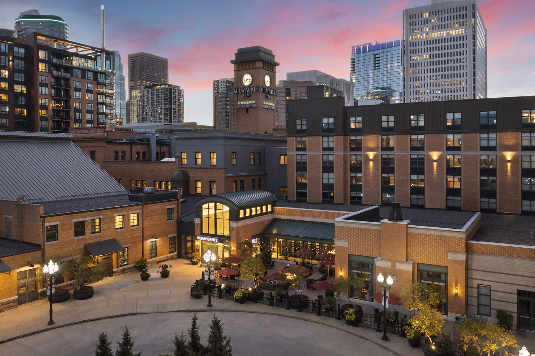A cityscape at sunset shows modern and historic buildings, a clock tower, outdoor dining area with tables and chairs, trees, and people enjoying the evening under a colorful sky.