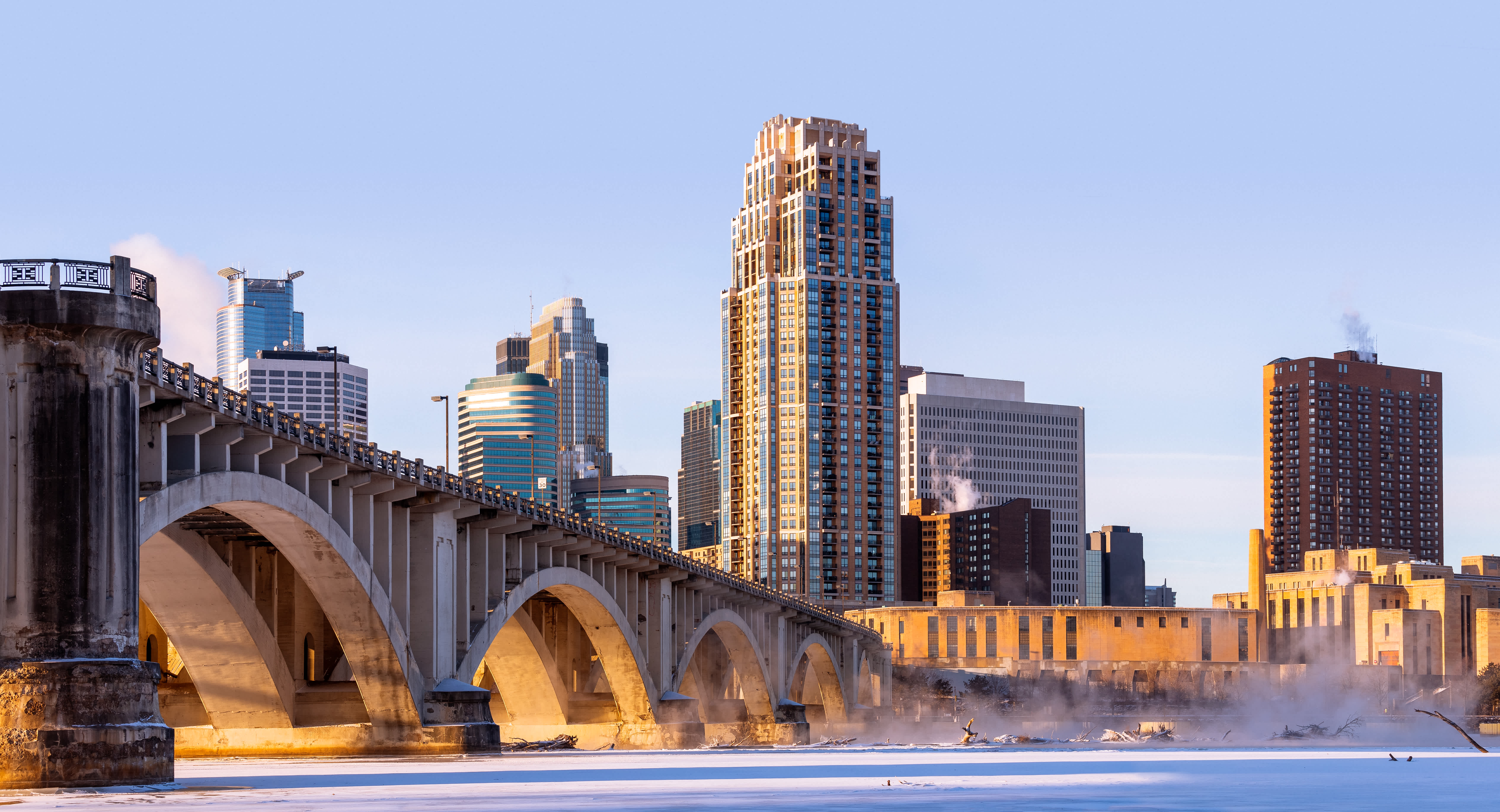 A city skyline with tall buildings, a large arched bridge, and smoke rising from rooftops on a cold winter morning with snow and ice along the river.